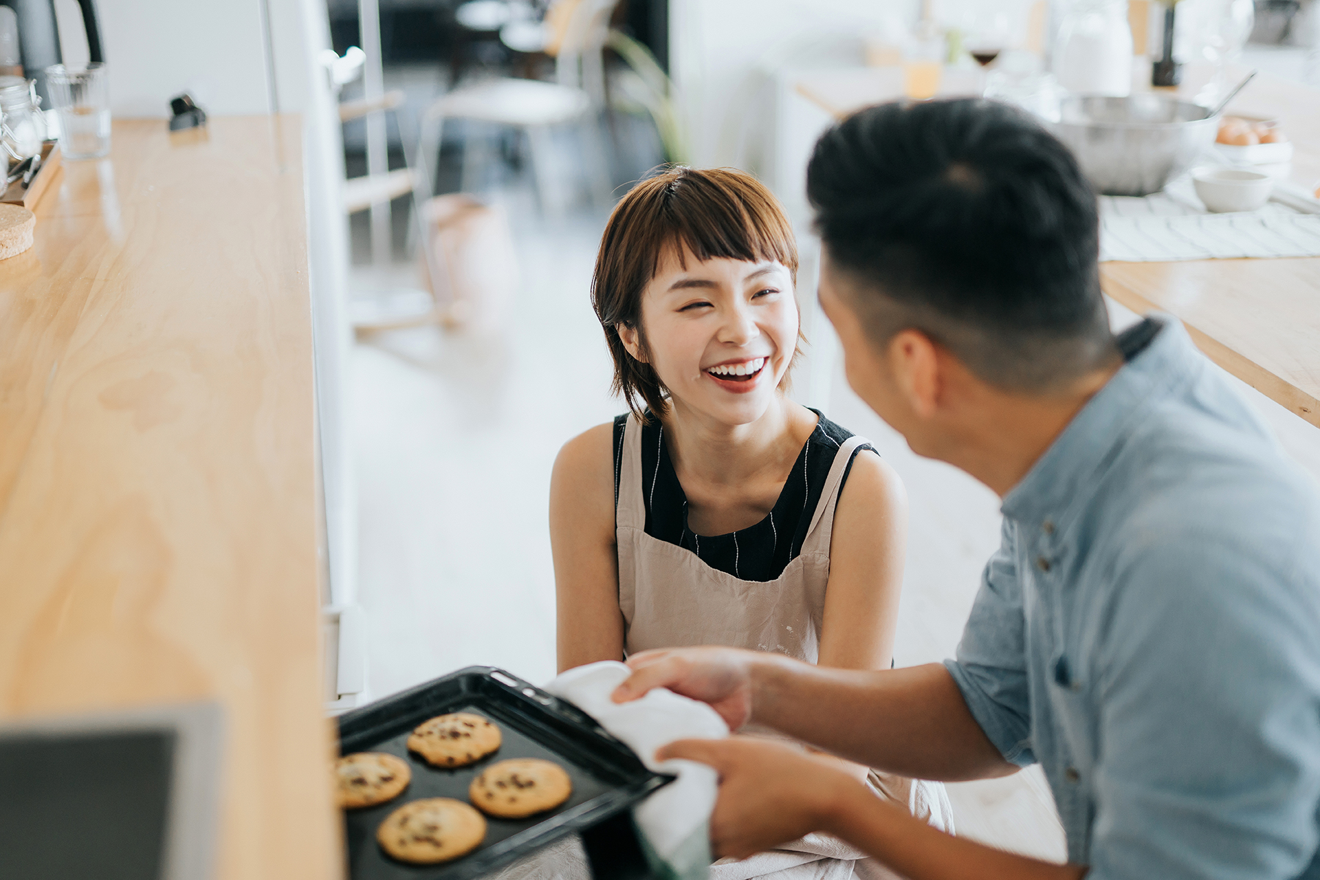 Joyful young Asian couple smiling at each other while taking out fresh home baked cookies from oven in a domestic kitchen