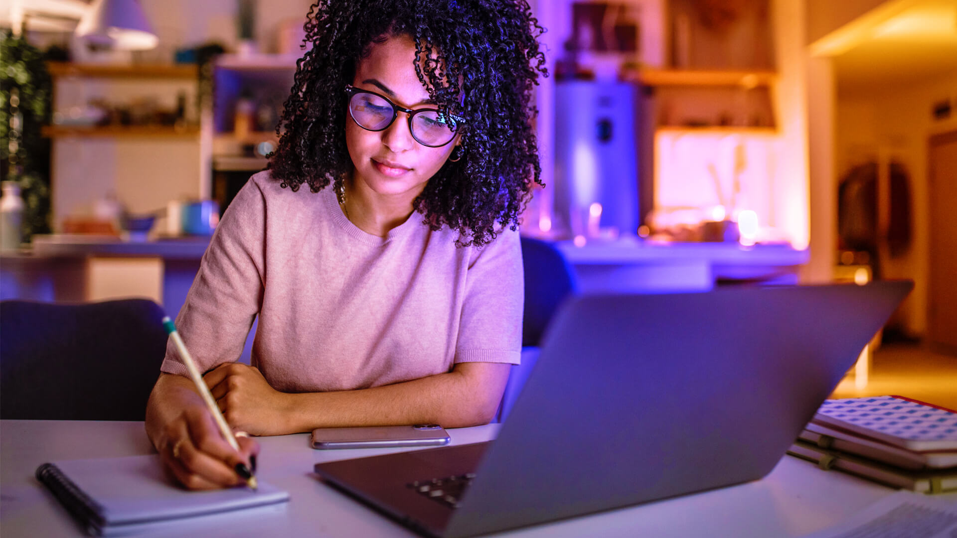 young woman sitting at a desk in front of an open laptop. she is writing in a notebook